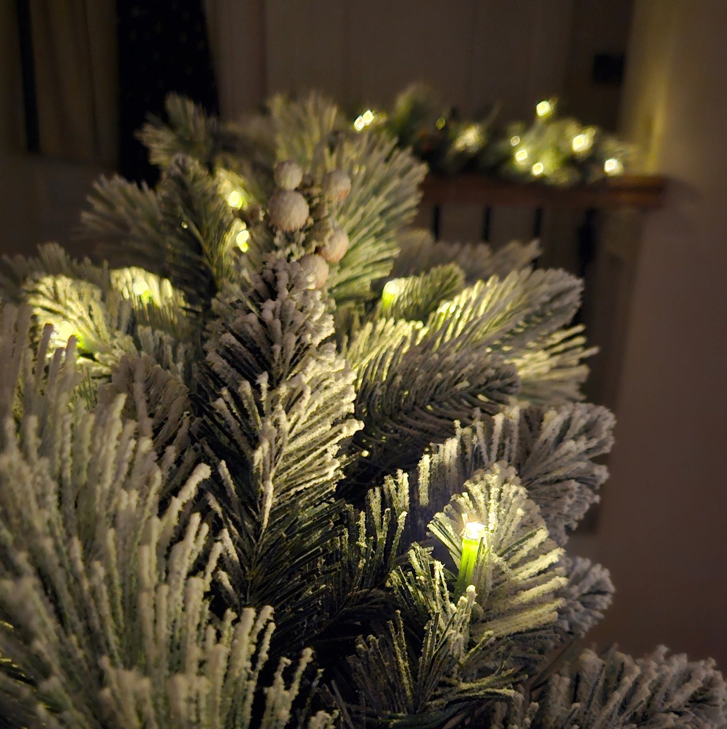 Close-up of a frosted artificial Christmas tree branch with warm white string lights glowing through the needles in a dim indoor setting.