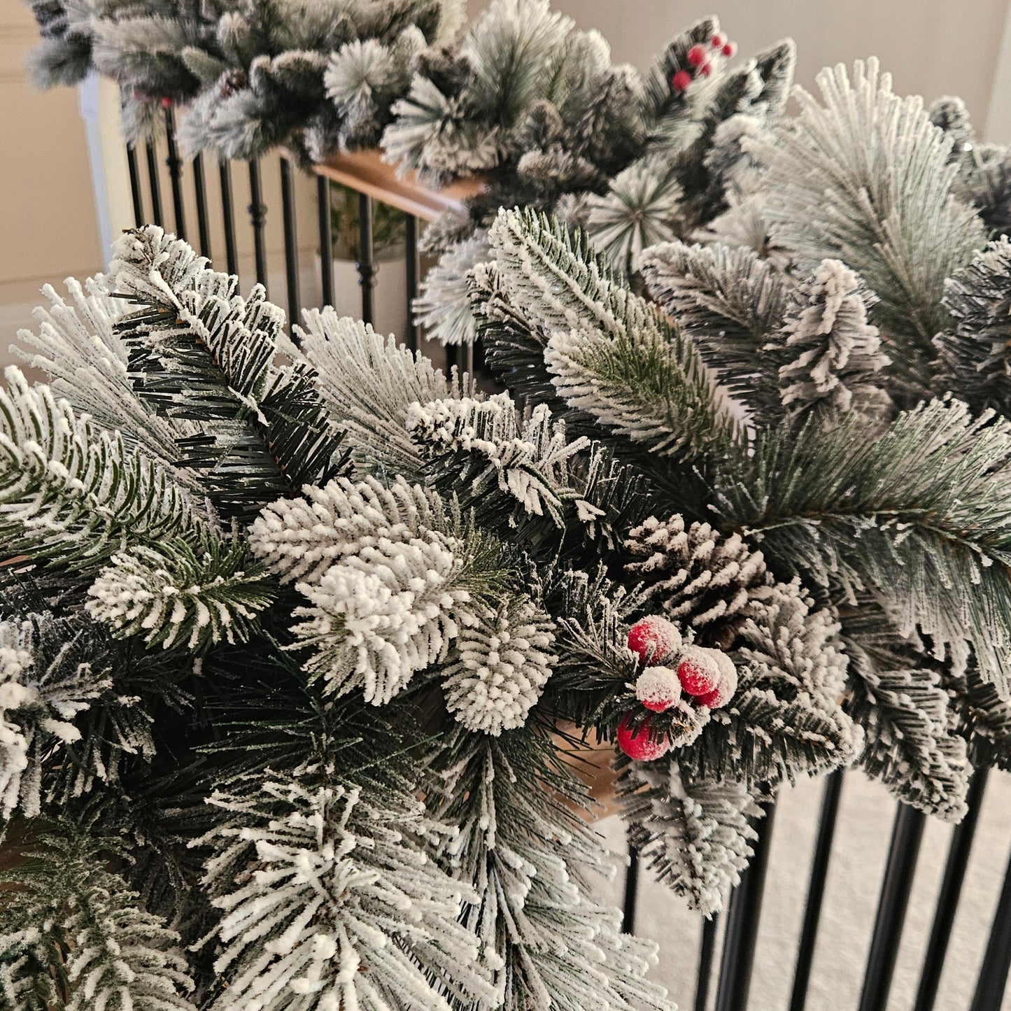 A close-up of a frosted Christmas garland with white snow-like dusting, pinecones, and small red berries hanging indoors near a staircase railing.