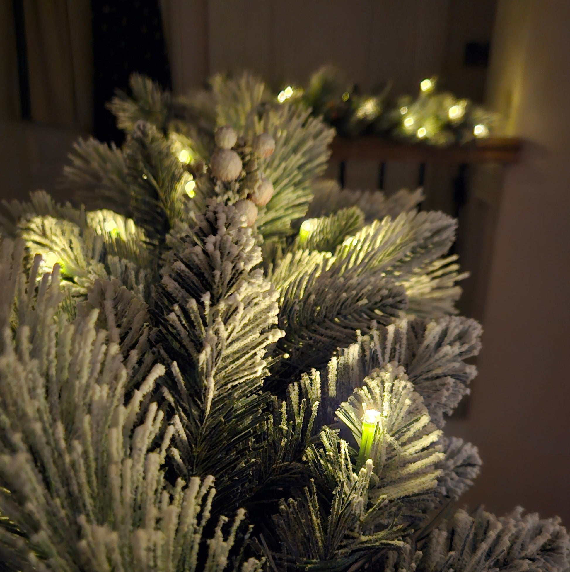 Close-up of a frosted artificial Christmas tree branch with warm white string lights glowing through the needles in a dim indoor setting.