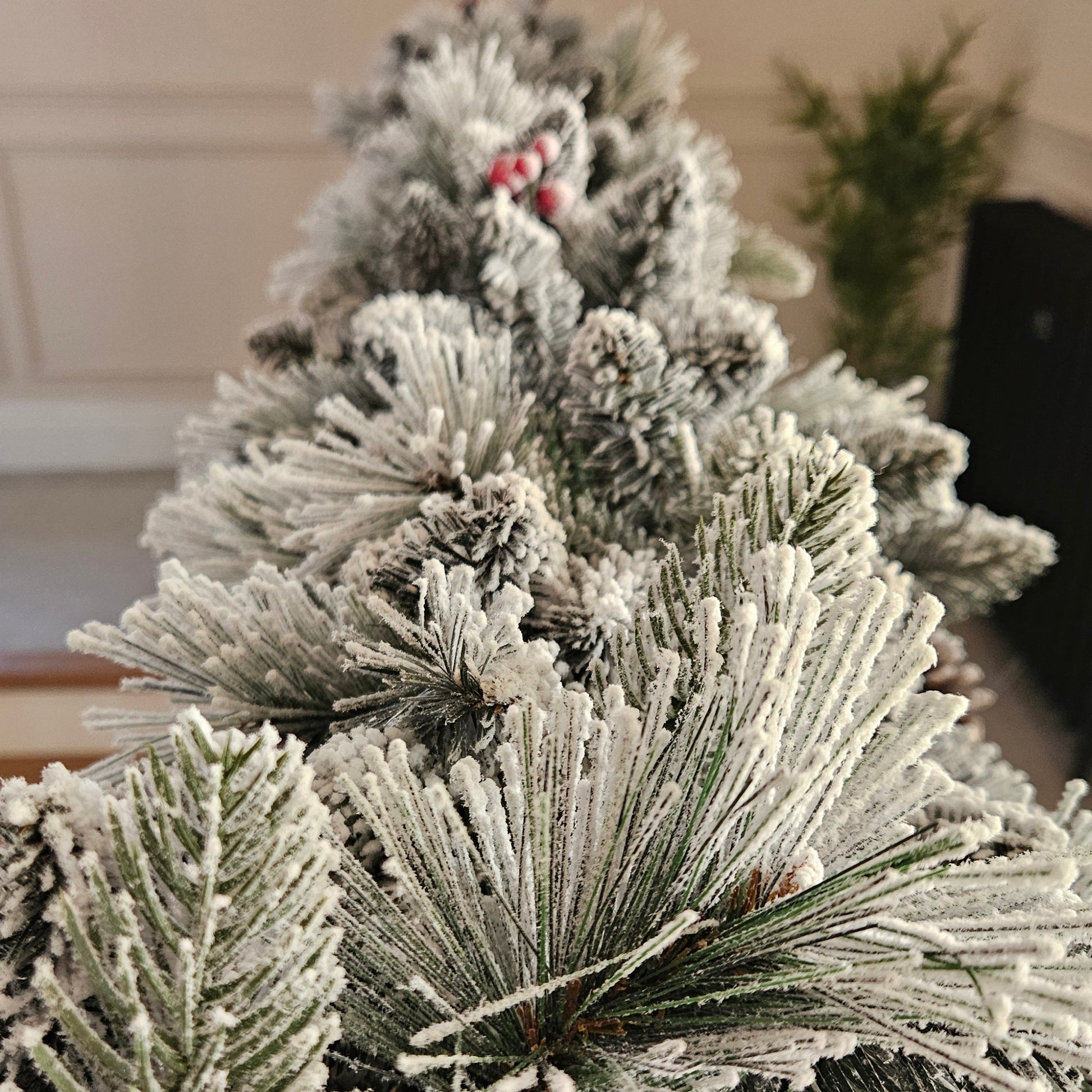 Close-up of a flocked artificial Christmas tree branch with white snow-like dusting, green needles, and small red berries, shown indoors against a softly blurred background.