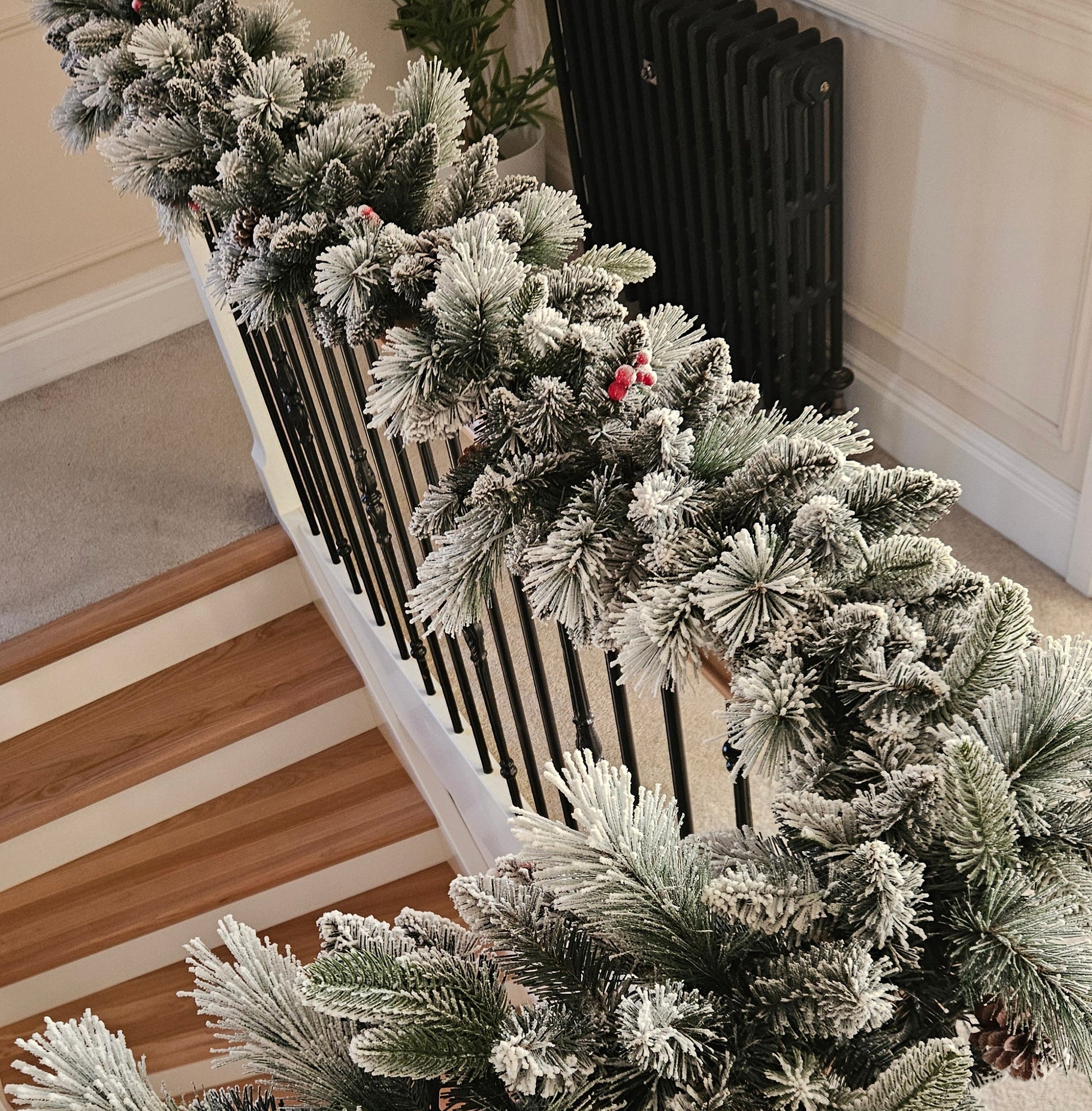 Frosted Christmas garland draped over a black metal staircase railing, decorated with small red berries, in an indoor staircase setting.